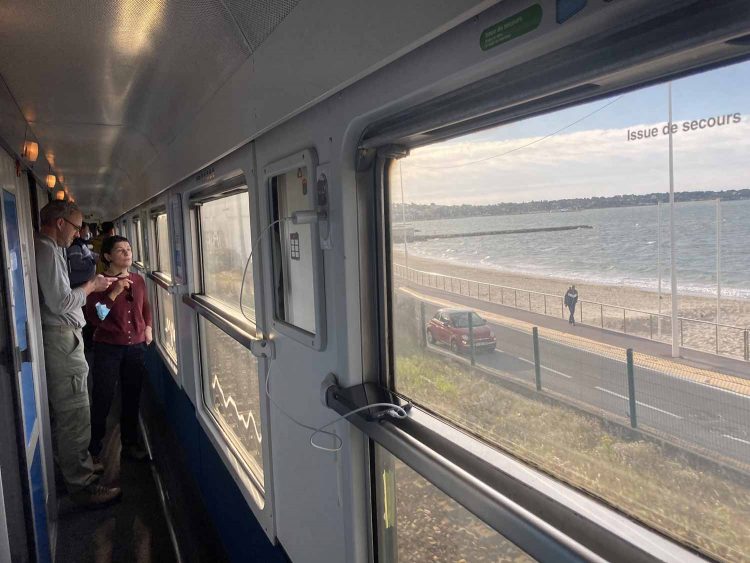 Dans l'allée d'un wagon, un couple de passagers regarde le paysage de bord de mer qui défile par la fenêtre.