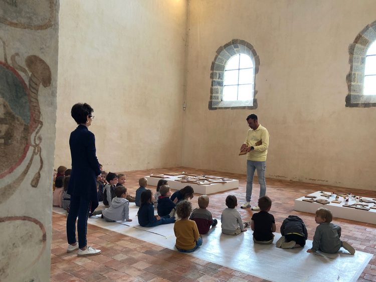 Dans l'ancien chœur de la chapelle, un homme debout présente des socles en bois à une classe d'écoliers assis devant lui, dans le cadre de l'exposition-atelier "la parole et le geste".