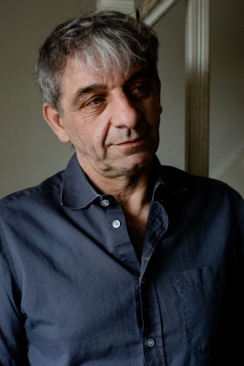 Portrait de face de Gilles C., climato-sceptique. Cheveux gris courts, yeux bleus, chemise sombre entrouverte.