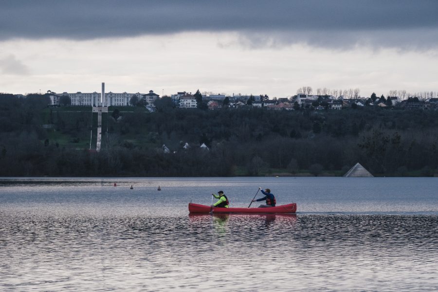 Deux kayakistes dans un kayak rouge sont en train de pagayer sur les étangs de l'île de loisirs de Cergy. En arrière-plan, l'Axe Majeur.