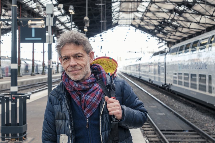 Sébastien, 58 ans, pose en tête de quai, à la gare de Lyon, visage souriant, sac sur l'épaule, prêt à partir !