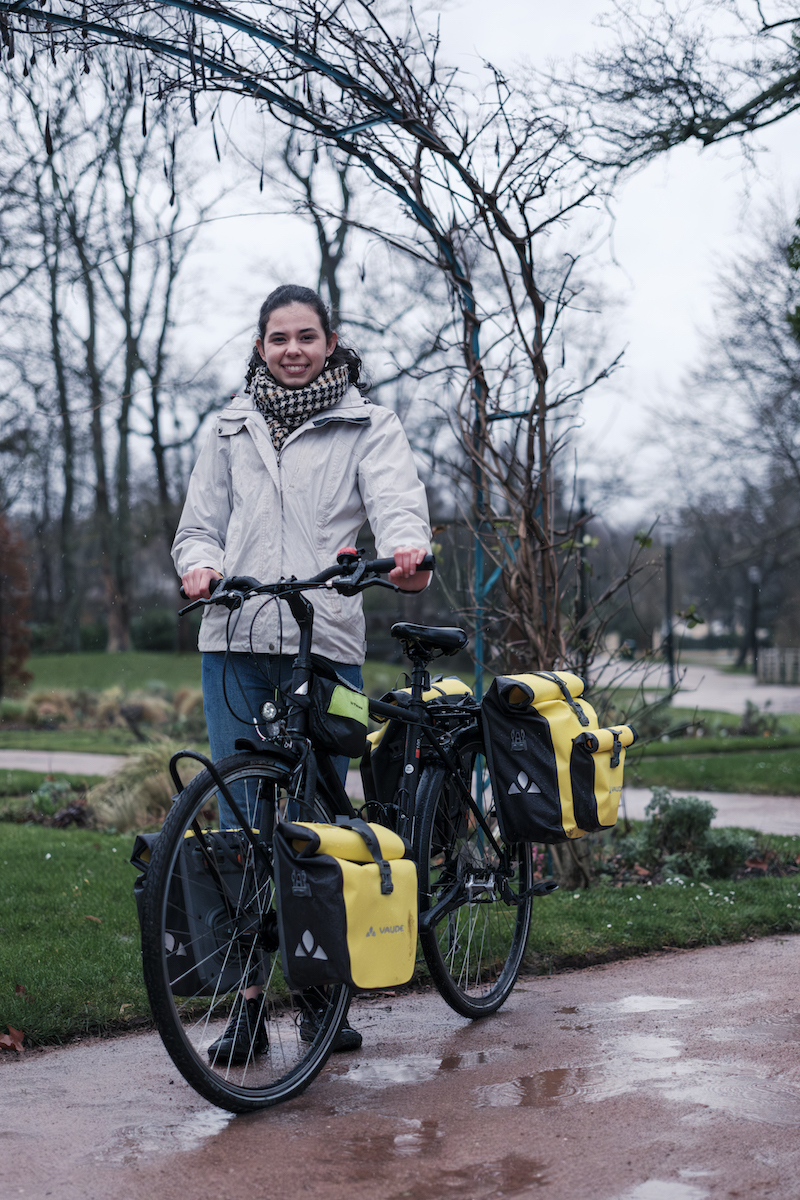 Dans un parc au Vésinet (Les Yvelines), le portrait de Lucie Reyx, tout sourire à côté de son vélo. Quatre sacoches jaunes; deux à l'avant et deux à l'arrière agrémente son vélo