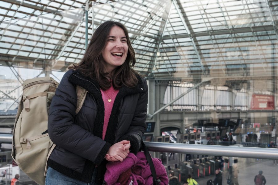 Clara, 24 ans, grande amatrice de voyages en train, pose à la gare de Lyon, sur une mezzanine surplombant le Hall 1, le visage illuminé d'un large sourire, sac sur le dos, prête pour le départ !
